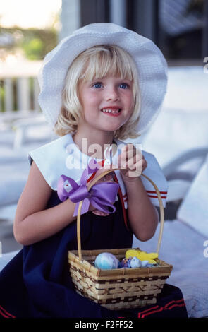 A young blonde American girl happily poses in her Easter dress and Easter hat with a basket of colored Easter eggs and candies on her lap. Dressing up and coloring eggs to celebrate the Easter holiday have long been traditions for children in the United States. Stock Photo