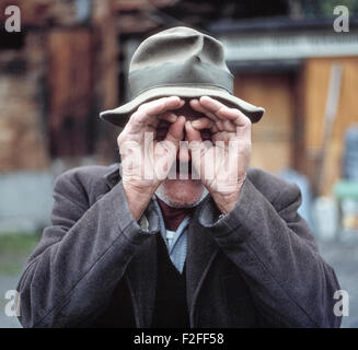 A grizzled old man in Switzerland mugs for the camera by putting his hands to his eyes to pretend he is looking through a pair of binoculars to spy on the photographer. Stock Photo