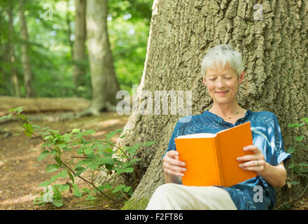 Smiling senior woman reading book against tree in woods Stock Photo