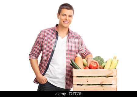 Young man leaning on a crate full of fresh vegetables and looking at the camera isolated on white background Stock Photo