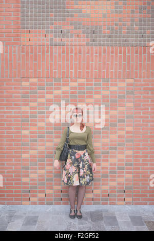 Young handsome redhead caucasian woman, leaning against a wall, looking in camera, smiling - pin up, carefreeness concept - wearing green shirt, floral skirt, bag and sunglasses Stock Photo