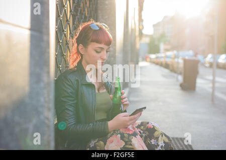 Young handsome caucasian redhead woman using smartphone holding a beer and using a smartphone, looking down and tapping the screen - social network, technology, communication concept - backlight Stock Photo
