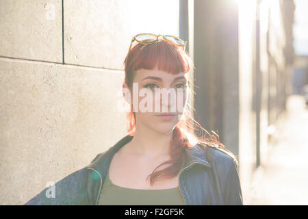 portrait of young handsome caucasian redhead woman leaning against a wall, looking in camera, serious - pensive concept - wearing leather jacket and green shirt - backlight Stock Photo