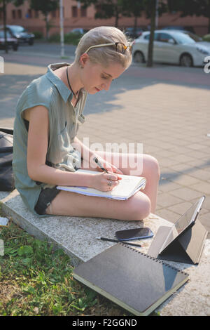 young handsome caucasian blonde italian designer sitting on a small wall, using smartphone and tablet and sketching on a notebook - creativity, enlightenment, working concept Stock Photo