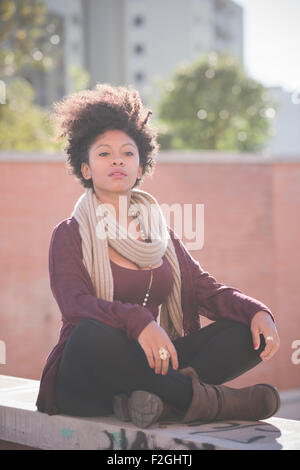 Young beautiful black curly hair african woman sitting on a small wall cross-legged, overlooking left, pensive - carefreeness, youth, pensive concept Stock Photo