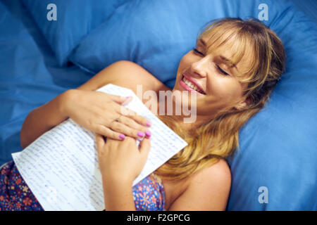 Young caucasian teenager receiving love letter from her new boyfriend. The girl lays on bed and holds the letter against her che Stock Photo