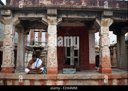 A hindu priest is reading on his smartphone in the Virupaksha temple. He is seated near the ( small)  temple of a hindu deity. Stock Photo