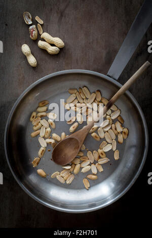 Roasted peanuts in steel skillet with wooden spatula on wooden tabletop with peanuts in shells. Top view Stock Photo