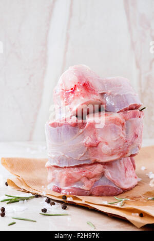 Stack of raw osso buco meat on crumpled paper with salt, pepper and rosemary over white marble as background Stock Photo