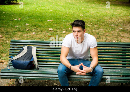 Young Male Student Sitting on the Bench in a Park, next to his Back Pack, While Thinking and Looking Away Seriously. Stock Photo
