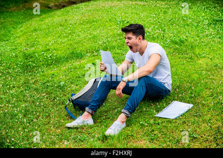 Young Bored, Tired Male Student Studying his Lessons while Yawning, Sitting on Grass in City Park, Smiling at Camera Stock Photo