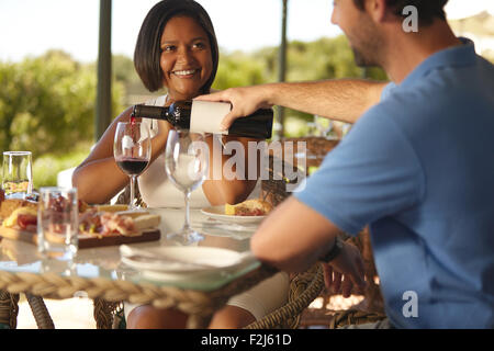 Happy young woman sitting at table with man pouring a glass of red wine at winery restaurant. Stock Photo