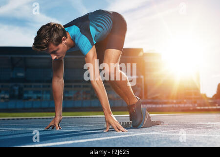 Young male runner taking ready to start position against bright sunlight. Sprinter on starting block of a racetrack in athletics