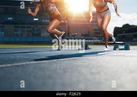 Sprinters starts out of the blocks on athletics racetrack with bright sunlight. Low section shot of female athletes starting a r Stock Photo