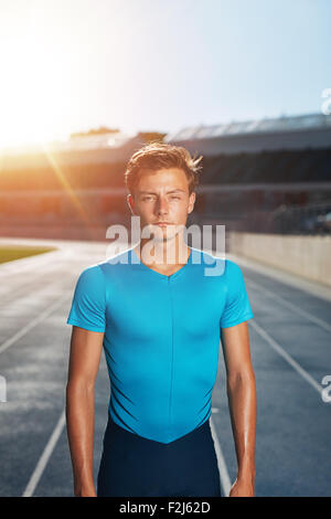 Young man after run standing on outdoor stadium race track. Fit male athlete looking at camera with sun flare. Stock Photo