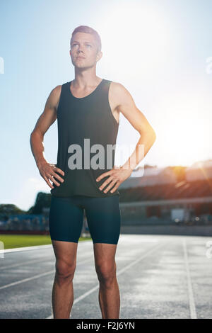 Fit young man standing with his hands on hips looking away. Determined male athlete preparing for a run on athletics stadium rac Stock Photo