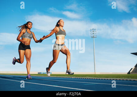 Young woman running a relay race and giving relay baton to her teammate. Female runner passing the relay baton during race. Stock Photo