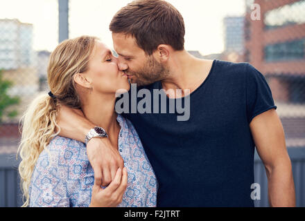 Amorous attractive young couple enjoy a romantic kiss as they stand arm in arm outdoors in an urban street