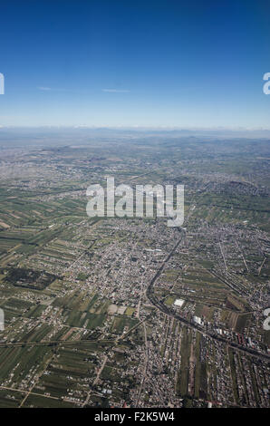 Iztapalapa Aerial View Urban Sprawl Agricultural Fields Mexico City Mexico | IZTAPALAPA, MEXICO CITY, Mexico — Agricultural fields and a dense settlement grid meet on the eastern fringe of Mexico City in this aerial view over Iztapalapa, where farmland gives way to expanding residential streets. A road corridor cuts diagonally through the pattern, tracing the edge of continued urban growth into the Valley of Mexico's agricultural periphery. Mountains and haze mark the horizon. Stock Photo