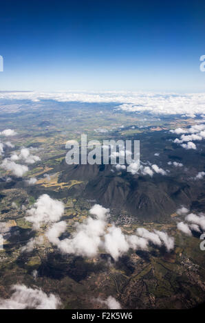 Mexico Aerial Landscape View From Airplane With Mountains Fields And Clouds | MEXICO — Clouds part over a mountain ridge and farmland in central Mexico, offering an airplane passenger's view of the country's volcanic highlands. Dark, forested slopes contrast with lighter cultivated fields below, a pattern typical of the Trans-Mexican Volcanic Belt that spans the nation's central plateau. A small town sits nestled at the base of the ridge, linked by roads threading through the valley. Stock Photo