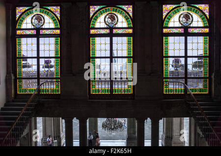 Chapultepec Castle Stained Glass Windows Mexico City | MEXICO CITY, Mexico — Three arched stained glass windows featuring the Mexican coat of arms illuminate a grand stone staircase inside Chapultepec Castle. Construction of the palatial structure began in 1785 under Viceroy Bernardo de Gálvez, eventually becoming the only royal castle in North America when it served as the residence for Emperor Maximilian I. The interior architectural detailing, including these vibrant leaded glass panels, integrates European neoclassical craftsmanship with nationalistic iconography symbolizing the Aztec foun Stock Photo