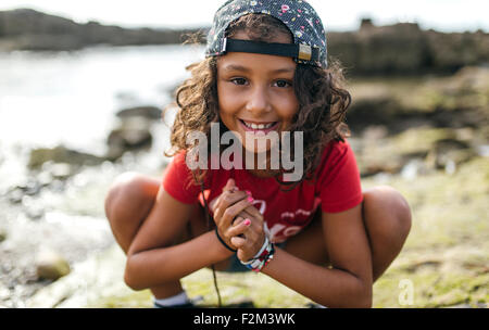 Spain, Gijon, portrait of smiling little girl crouching at rocky coast Stock Photo