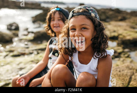 Spain, Gijon, portrait of laughing little girl and her friend in the background sitting at rocky coast Stock Photo