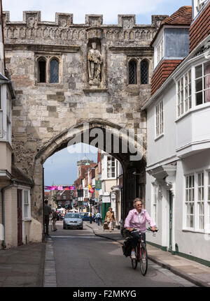 Older woman rides bicycle in Cathedral Close, Salisbury, England. Crenellated stone city wall with gothic arch & new town beyond Stock Photo