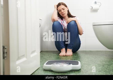 Unhappy Teenage Girl Sitting On Floor Looking At Bathroom Scales Stock Photo