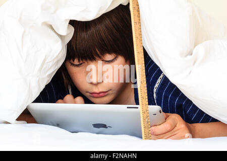 Caucasian child, boy, 10 - 11 year old. Half hiding under white duvet with a ruler propping it up. Head and shoulders of boy as he uses ipad, Stock Photo