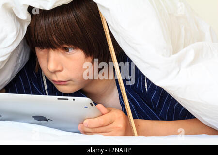 Caucasian child, boy, 10 - 11 year old. Half hiding under white duvet with a ruler propping it up. Head and shoulders of boy as he uses ipad, Stock Photo