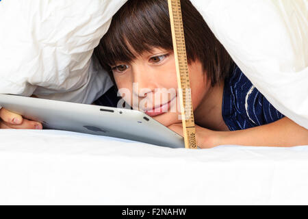 Caucasian child, boy, 10 - 11 year old. Half hiding under white duvet with a ruler propping it up. Head and shoulders of boy as he uses ipad, Stock Photo