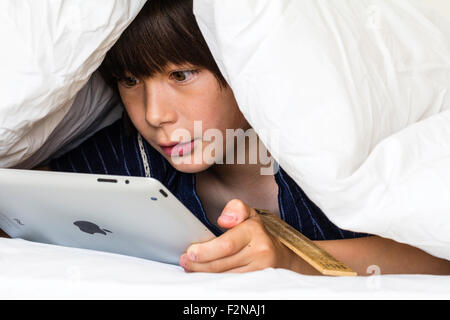 Caucasian child, boy, 10 - 11 year old. Half hiding under white duvet with a with face peeking out. Head and shoulders of boy as he uses ipad, Stock Photo