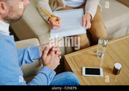 Male sitting in armchair while psychiatrist making notes in medical document Stock Photo