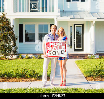 Portrait of a smiling young couple by sold sign standing against sky ...