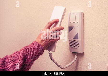 a mature woman is picking up an intercom telephone receiver from a wall-mounted unit. Stock Photo