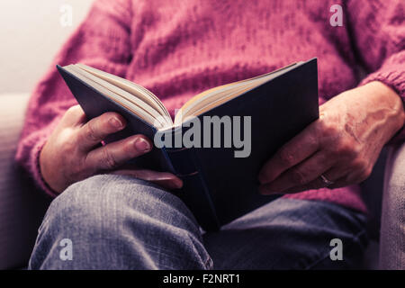 a mature person is sitting in an armchair, reading a book. Stock Photo