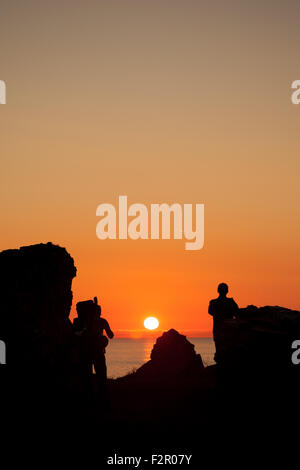 2 men standing between rocks silhouetted against an Orange sunset Stock Photo