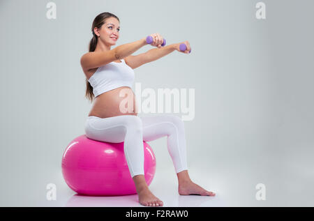 Portrait of a pregnant woman doing exercises with dumbbells on a fitness ball isolated on a white background Stock Photo