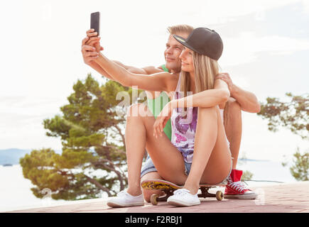 Happy couple with sitting outdoors on skateboard and taking a selfie Stock Photo