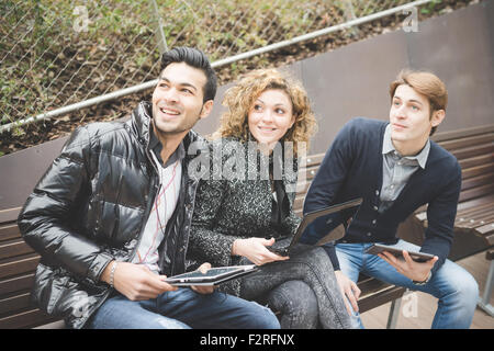 Multiracial business people working outdoor in town sitting on a bench, connected with technological devices like tablet and laptop, overlooking right, smiling - business, work, carefreeness concept Stock Photo