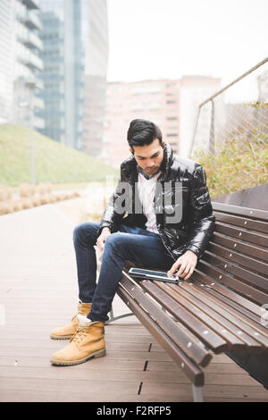 Young handsome indian contemporary businessman seated on a bench in a park using tablet while listening music with earphones looking downward on the screen  - business, work, study, technology concept Stock Photo