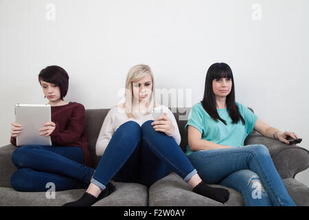 Mother and her two daughters sitting on a sofa watching TV , using an ipad and iphone. Stock Photo