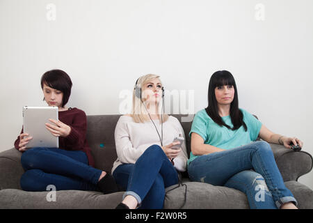 Mother and her two daughters sitting on a sofa watching TV , using an ipad and listening to music. Stock Photo