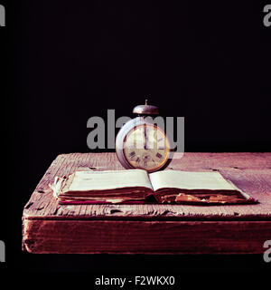 an old alarm clock and an open old book on a rustic wooden table, against a black background Stock Photo