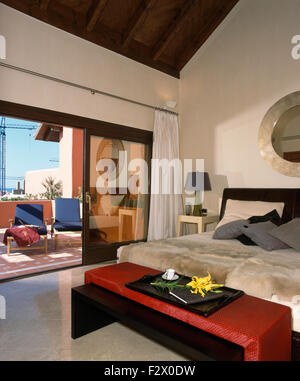 Red stool below bed with faux fur throw in modern Spanish bedroom with open glass doors to patio Stock Photo