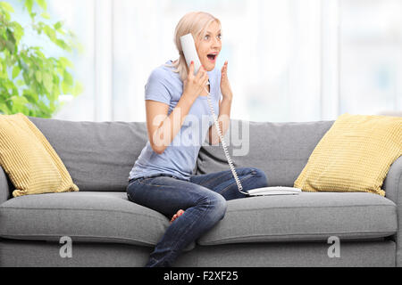 Surprised blond woman talking on the telephone and gesturing happiness seated on a couch at home Stock Photo