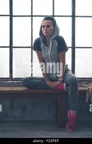 Pensive, a woman pauses, sitting on a loft gym bench, leaning her hands against her foot as she stretches and thinks about the workout she has just done. Stock Photo