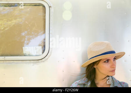 A young woman wearing a hat sitting in the shade of a silver coloured trailer. Stock Photo