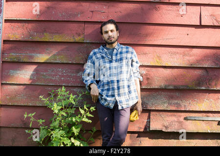 Bearded man wearing a checkered shirt leaning against a wooden wall in the shade. Stock Photo